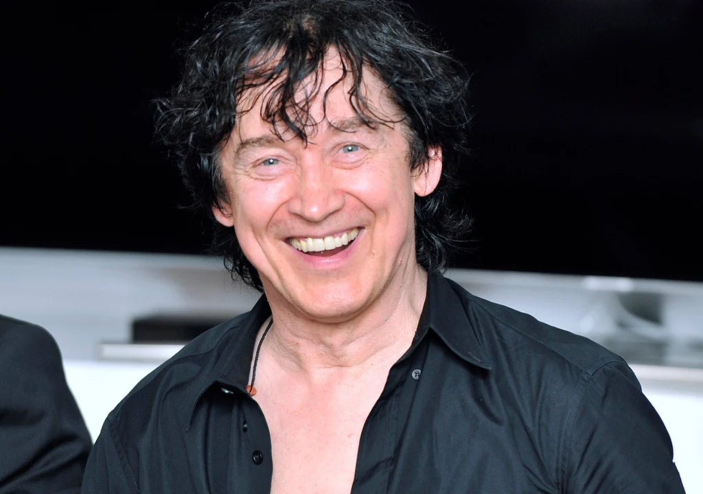 Jan Borysewicz Uśmiechnięty mężczyzna o ciemnych, lekko potarganych włosach w ciemnej, rozpiętej koszuli, stoi na tle rozmazanego wnętrza.