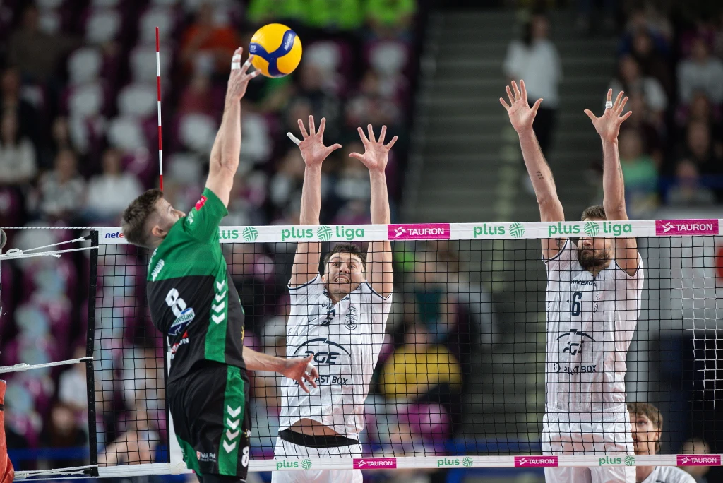 Kadr z meczu PGE Projekt Warszawa - ChKS Chełm Zawodnik w zielonym stroju atakuje piłką siatkową na boisku, podczas gdy dwóch zawodników w białych strojach próbuje zablokować piłkę przy siatce w hali sportowej.