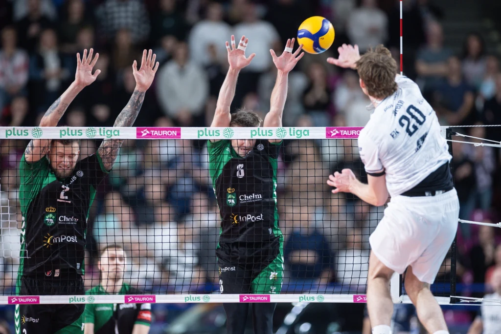 Atakuje Linus Weber z PGE Projektu Warszawa Zawodnik w białym stroju atakuje piłkę siatkową ponad siatką, podczas gdy dwóch przeciwników w zielono-czarnych strojach próbuje zablokować piłkę, a w tle widoczna jest publiczność.