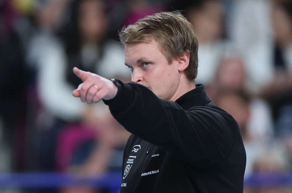 Tommi Tilikainen, nowy trener PGE Projektu Warszawa Mężczyzna w czarnej bluzie energicznie wskazuje palcem przed siebie, koncentrując wzrok w kierunku, w którym pokazuje, wokół niego rozmazane postacie ludzi siedzących na trybunach.