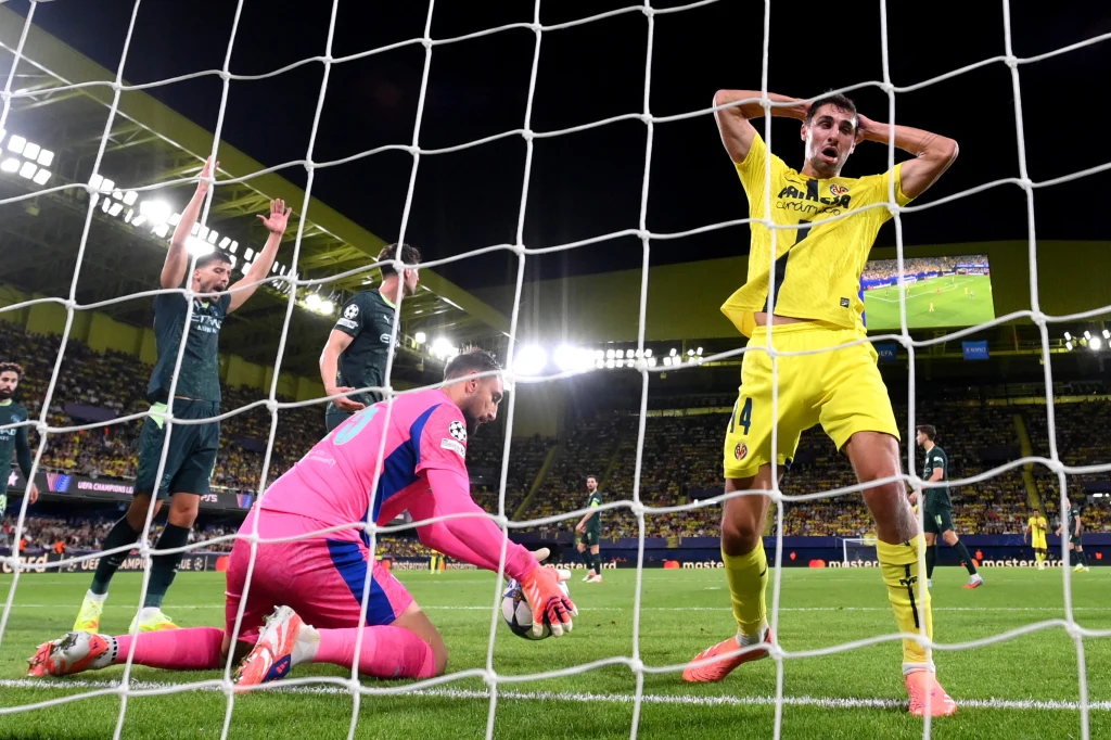 Kadr z meczu Manchester City - Villarreal Piłkarz w żółtym stroju z grymasem rozczarowania trzyma ręce na głowie, podczas gdy bramkarz w różowo-niebieskim stroju chwyta piłkę na linii bramkowej, obok niego widoczni są kolejni zawodnicy, a wszystko dzieje się na tle stadionowych trybun i pod sz...