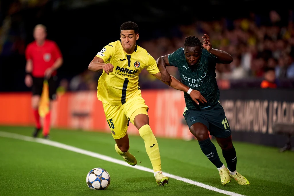 Kadr z meczu Manchester City - Villarreal Dwóch piłkarzy walczy o piłkę na boisku podczas meczu, jeden ubrany w żółty strój, drugi w ciemnozielony, obaj w dynamicznym ruchu tuż przy linii bocznej.