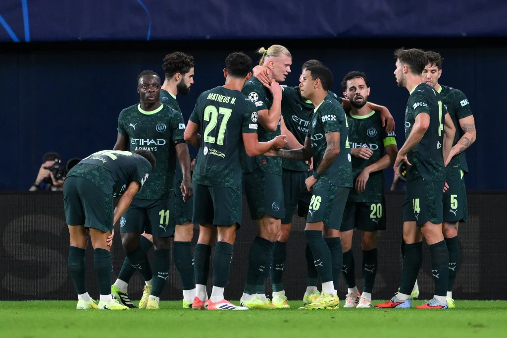 Kadr z meczu Manchester City - Villarreal Piłkarze drużyny Manchester City w zielonych strojach zebrani na boisku podczas meczu, kilku zawodników prowadzi rozmowę, widoczne loga sponsorów i numery na koszulkach.