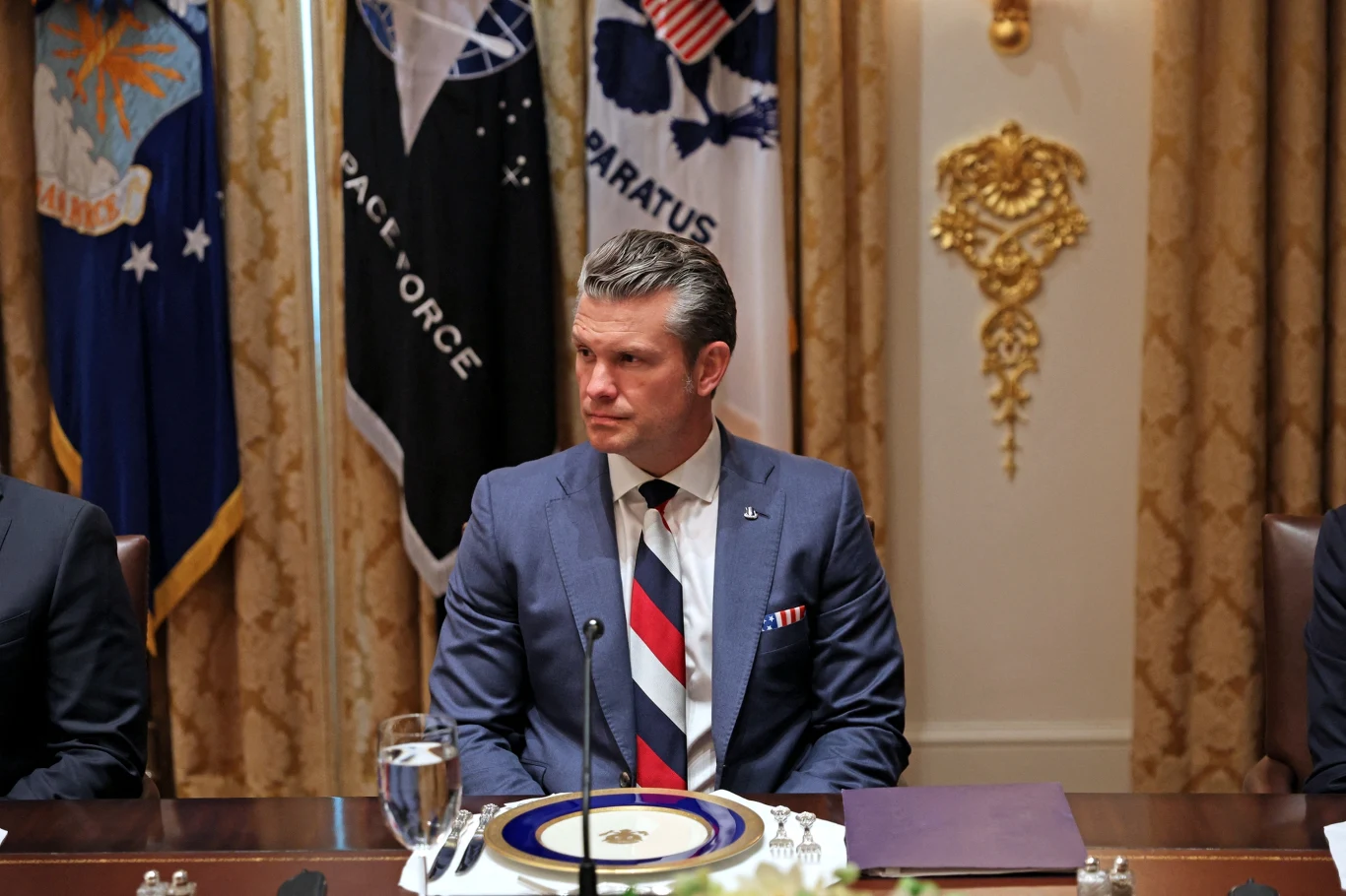Szef Pentagonu Pete Hegseth Mężczyzna w garniturze siedzi przy elegancko nakrytym stole konferencyjnym, na tle flag oraz ozdobnej ściany, przed nim znajduje się talerz i szklanka z wodą.