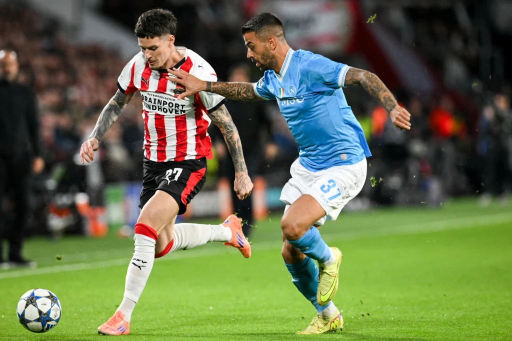 Liga Mistrzów: PSV Eindhoven ograło Napoli 6:2 Dwóch piłkarzy walczy o piłkę podczas meczu piłki nożnej, jeden w czerwono-białym stroju PSV Eindhoven, drugi w niebieskim stroju przeciwnika, z widoczną dynamiką i skupieniem na grze, w tle rozmazani kibice.