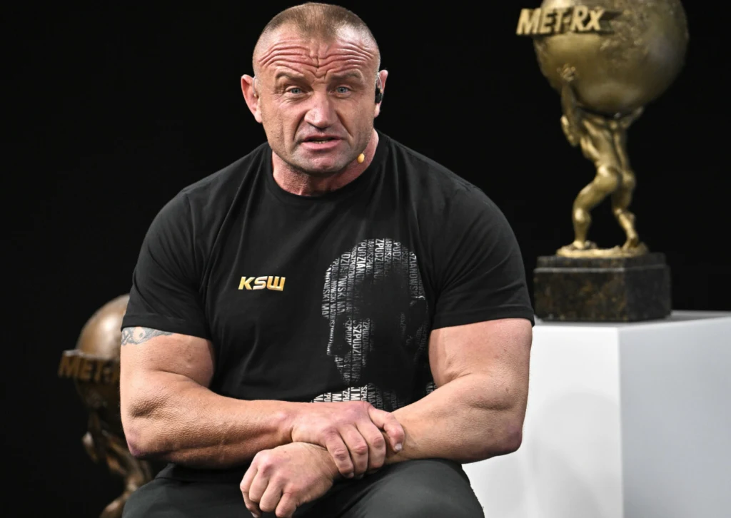 Muskularny mężczyzna w czarnej koszulce z nadrukiem, siedzący na tle dużych trofeów sportowych w kształcie człowieka niosącego kulę, atmosfera sceny wydaje się oficjalna i związana z wydarzeniem sportowym.