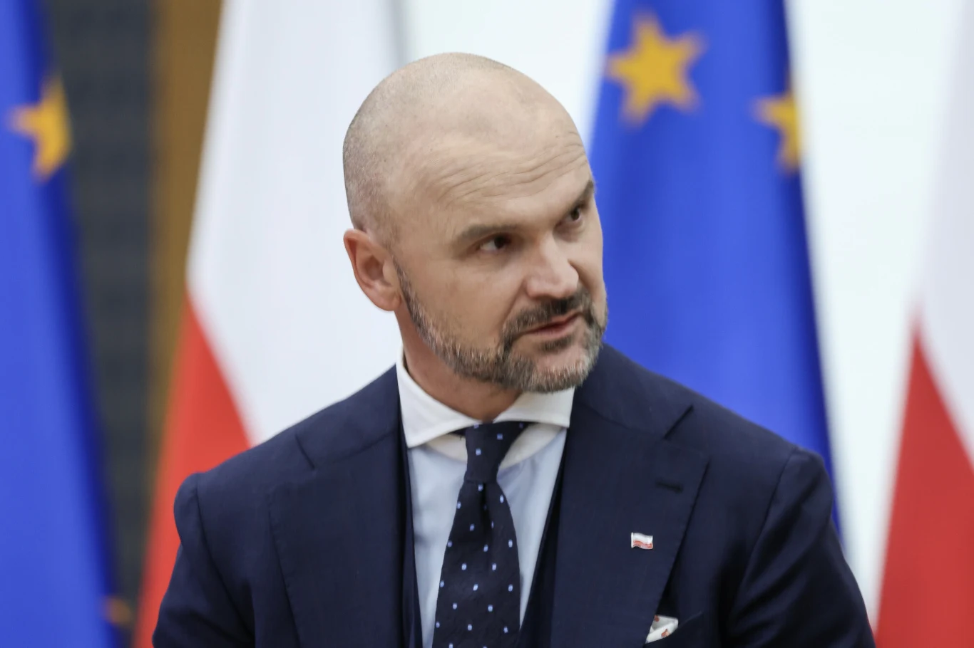 Rafał Brzoska ma pomysły, jak zaangażować firmy w budowanie bezpieczeństwa państwa Mężczyzna w garniturze z przypiętą miniaturową polską flagą na klapie, w tle widoczne flagi Polski oraz Unii Europejskiej.