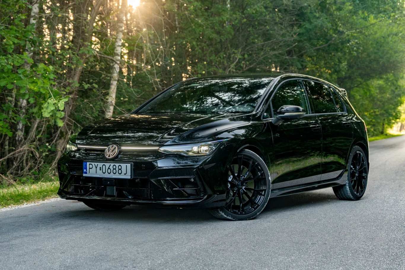 Volkswagen Golf R Black Edition wyróżnia się czarnymi dodatkami, ale wbrew pozorom całe nadwozie nie musi być czarne Czarny, sportowy Volkswagen Golf stoi zaparkowany na drodze otoczonej zielonym lasem, oświetlony promieniami słońca przebijającymi się przez drzewa, widoczny jest numer rejestracyjny auta.