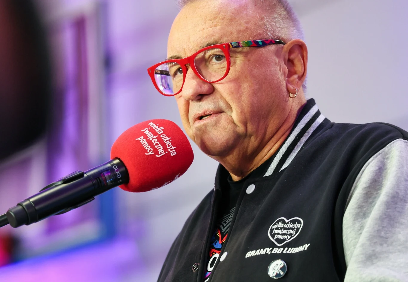 Mężczyzna w kolorowych okularach i czarnej kurtce z naszywkami, stojący przy czerwonym mikrofonie z logo Polskiego Radia, tło rozmyte, w odcieniach fioletowych.