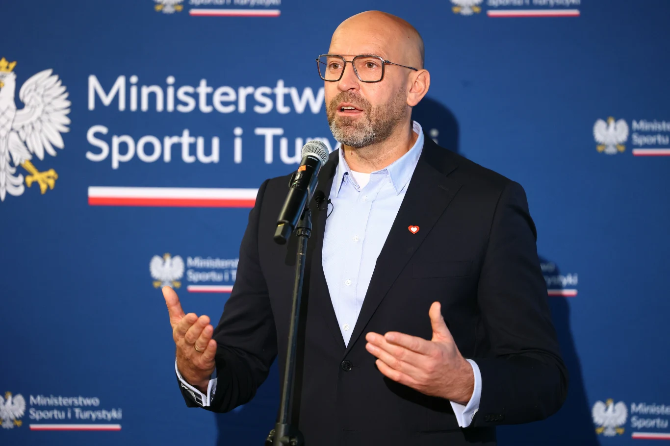 Mężczyzna w garniturze i okularach przemawia do mikrofonu na tle ścianki z logotypem Ministerstwa Sportu i Turystyki oraz godłem Polski.