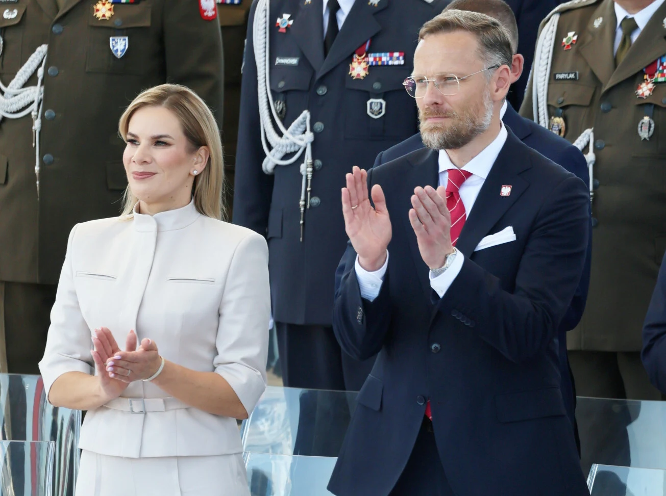 Szef kancelarii prezydenta RP uważa, że Pierwsza Dama powinna mieć "godne zarobki" Marta Nawrocka i mężczyzna w garniturze klaszczą, za nimi żołnierze na tle flag i odznaczeń.