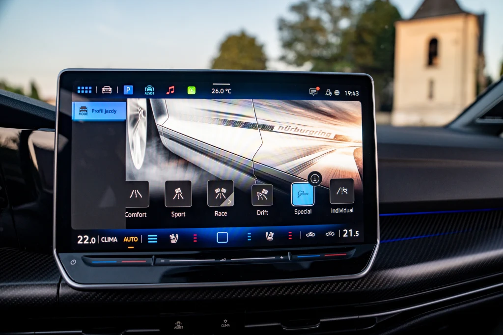 Golf R Black Edition seryjnie oferuje dodatkowe sportowe tryby jazdy - Special oraz Drift Dotykowy ekran systemu multimedialnego w samochodzie wyświetla opcje zmiany trybu jazdy, tło z rozmazanym widokiem zewnętrznym oraz fragment wnętrza pojazdu.