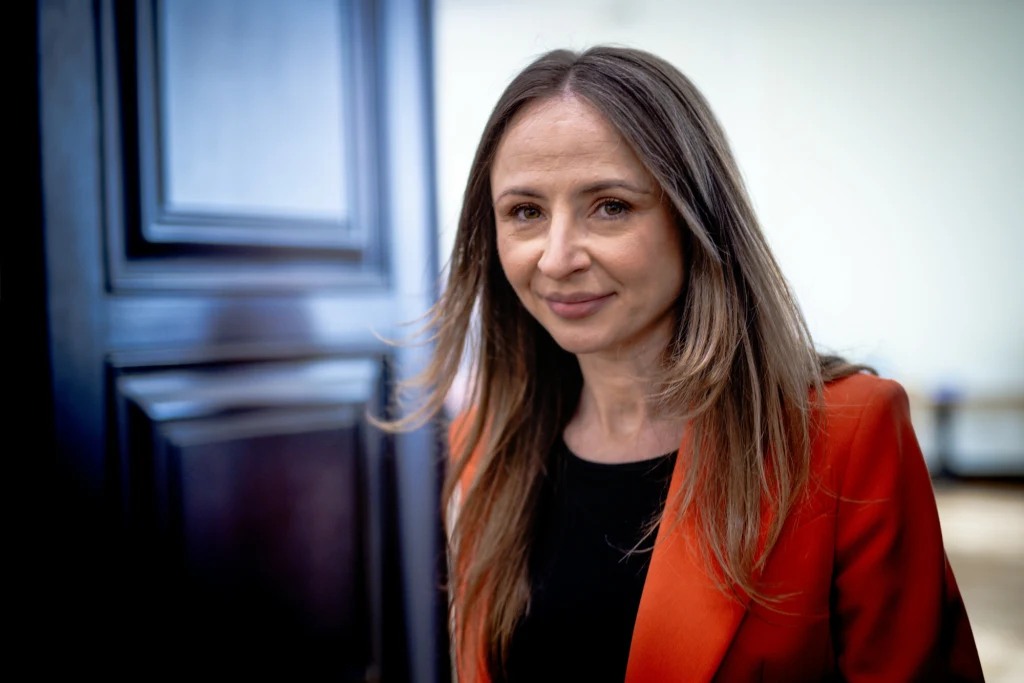 Kobieta o długich, jasnobrązowych włosach, ubrana w pomarańczową marynarkę i czarną bluzkę, stoi przy dużych, ciemnych drzwiach, patrząc łagodnie w stronę aparatu. Jasne tło kontrastuje z jej ubiorem.