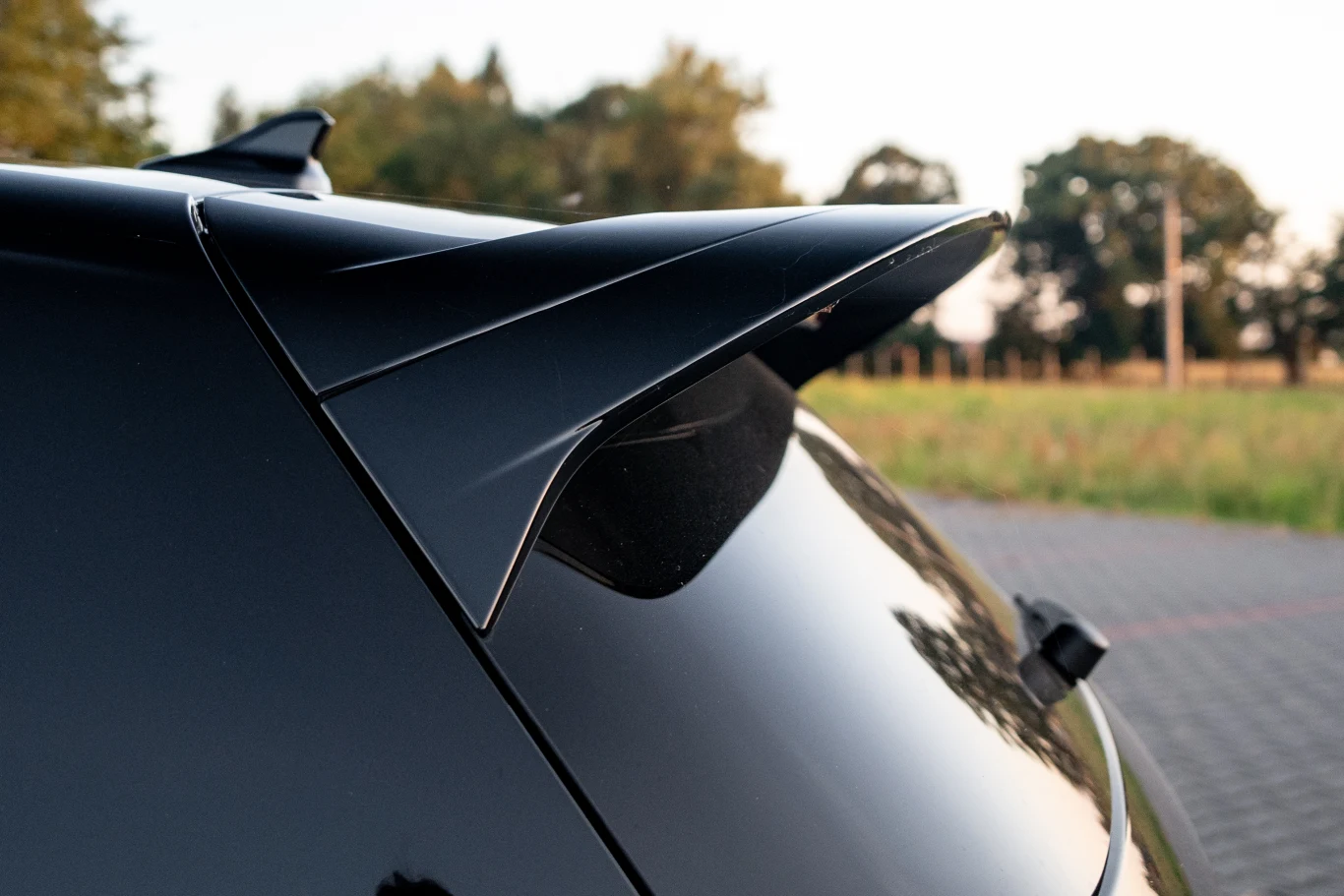 Volkswagen Golf R Black Edition standardowo ma powiększoną lotkę nad tylną szybą Czarny spoiler zamontowany na tylnej klapie samochodu, odbijający światło dzienne, w tle widoczne rozmyte drzewa oraz fragment parkingu.