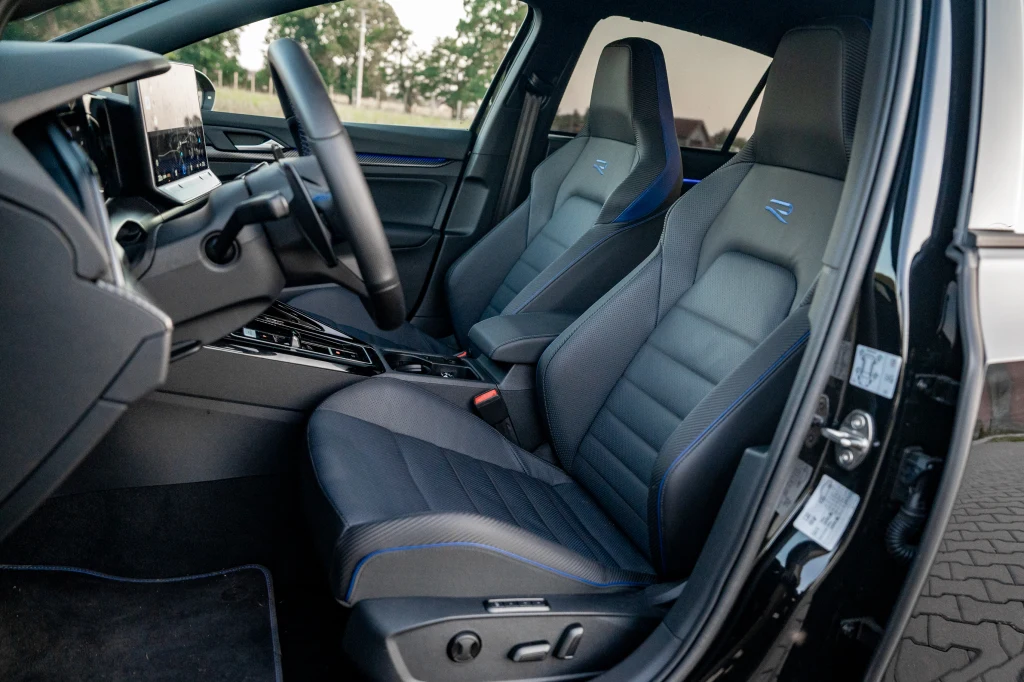 Golf R ma bardzo wygodne i dobrze trzymające w zakrętach fotele, ale ich siedziska mogłyby być dłuższe Wnętrze nowoczesnego samochodu z widoczną deską rozdzielczą, kierownicą oraz dwoma sportowymi fotelami z przeszyciami i oznaczeniem R na zagłówkach.