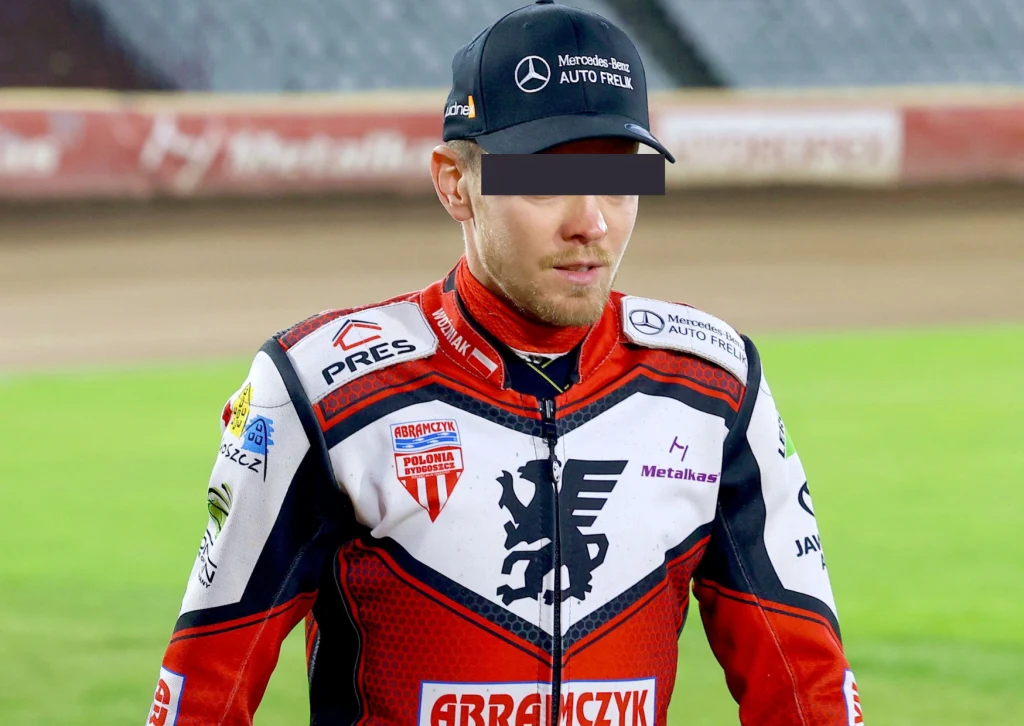 Szymon W. zatrzymany. Żużlowiec w profesjonalnym kombinezonie sportowym z widocznymi logotypami sponsorów stoi na stadionie żużlowym, w tle trybuny oraz tor.