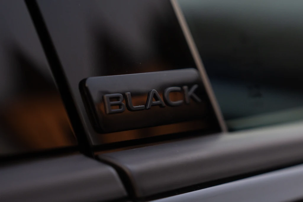 Czarny emblemat z czarnym napisem "BLACK" umieszczony na słupku B to jedyne miejsce, gdzie znajdziemy potwierdzenie, że faktycznie mamy do czynienia z Golfem R Black Edition Metaliczny emblemat z napisem BLACK umieszczony na czarnej powierzchni, prawdopodobnie na ramie drzwi pojazdu, z wyraźnym połyskiem lakieru.