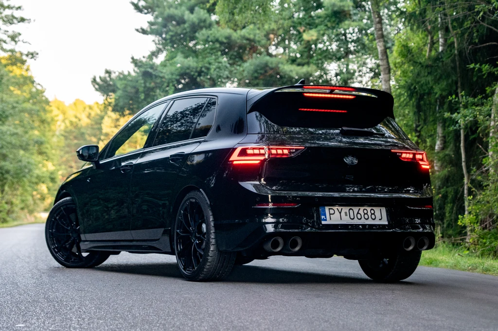 Volkswagen Golf R prowadzi się doskonale, a dzięki adaptacyjnemu zawieszeniu z 15 ustawieniami twardości, każdy znajdzie odpowiadające mu nastawy Czarny samochód osobowy stojący na asfaltowej drodze w otoczeniu zielonych drzew, auto wyróżnia się sportową sylwetką, nowoczesnymi światłami tylnymi oraz przyciemnianymi szybami.