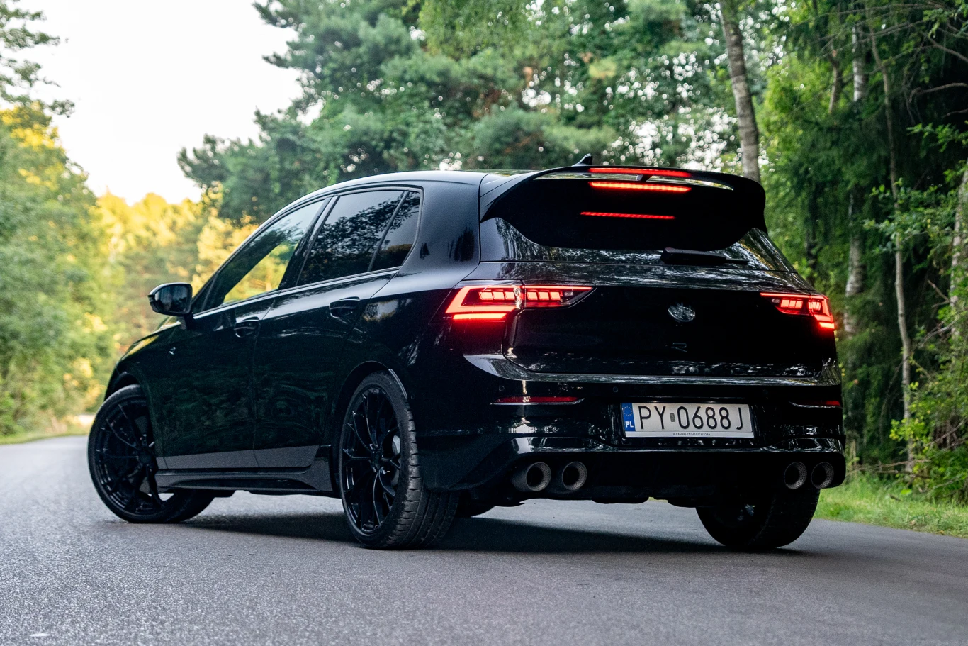Volkswagen Golf R prowadzi się doskonale, a dzięki adaptacyjnemu zawieszeniu z 15 ustawieniami twardości, każdy znajdzie odpowiadające mu nastawy Czarny samochód osobowy stojący na asfaltowej drodze w otoczeniu zielonych drzew, auto wyróżnia się sportową sylwetką, nowoczesnymi światłami tylnymi oraz przyciemnianymi szybami.