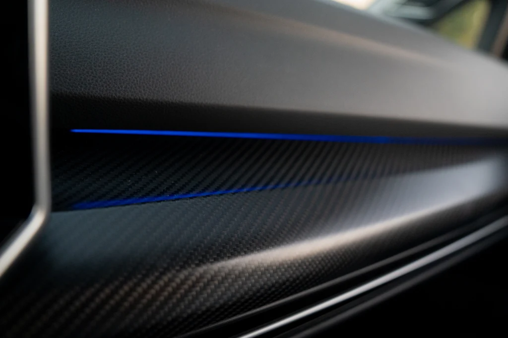 Ozdobne wstawki co prawda tylko imitują karbon, ale robią to naprawdę doskonale Element wnętrza samochodu wykonany z materiału imitującego włókno węglowe z niebieskim podświetleniem biegnącym wzdłuż powierzchni.