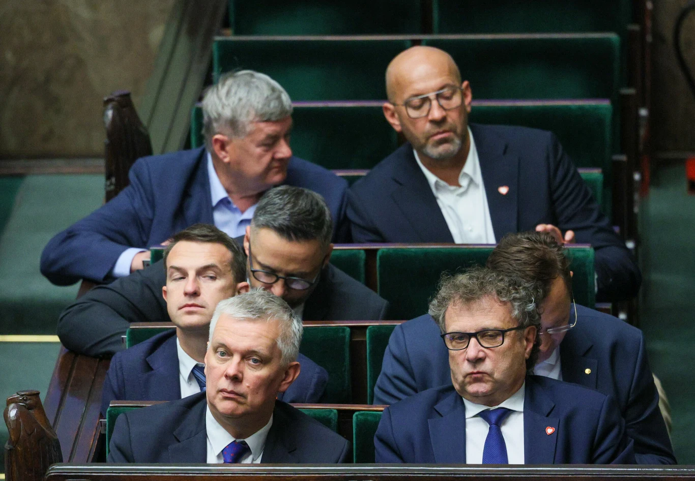 Grupa mężczyzn w garniturach siedzi w ławach parlamentarnych, skupieni na przebiegu obrad, niektórzy z nich patrzą w stronę mównicy, część rozmawia ze sobą.