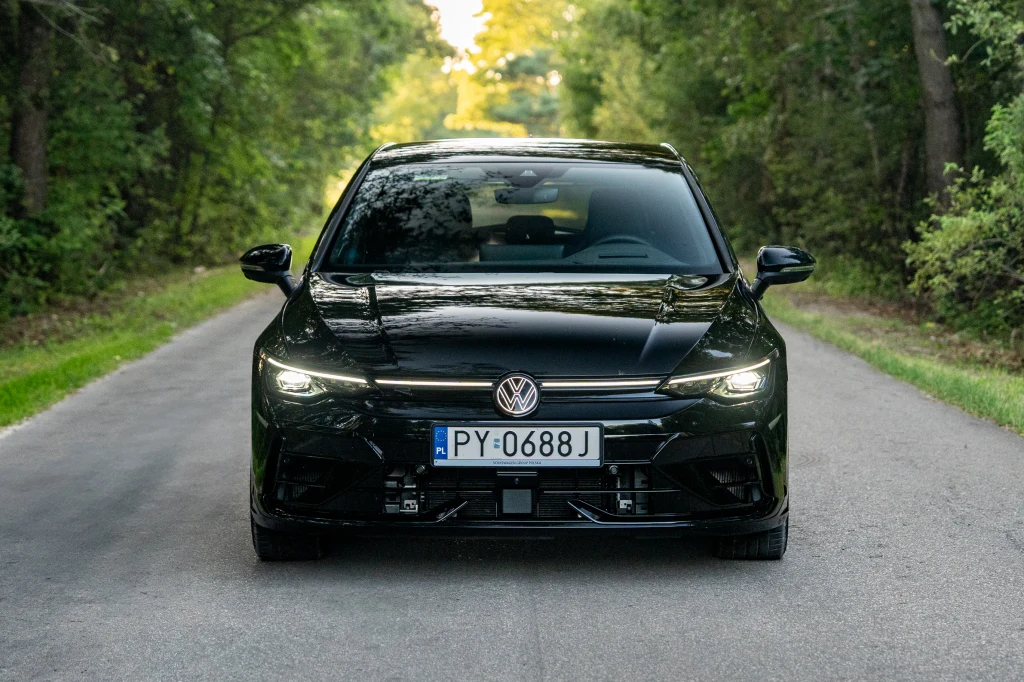 Zmodernizowanego Volkswagena Golf najłatwiej rozpoznać po świecącym się logo Czarny samochód marki Volkswagen stoi na środku asfaltowej drogi otoczonej gęstą zielenią po obu stronach, tablica rejestracyjna widoczna z przodu pojazdu, w tle jasny fragment drogi oświetlony słońcem.
