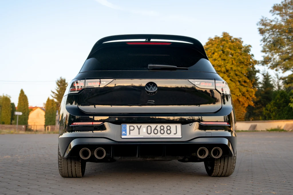 Volkswagen Golf R Black Edition Czarny samochód marki Volkswagen Golf R widziany z tyłu na tle jasnego nieba oraz zielonych drzew, z widocznymi czterema końcówkami wydechu i tablicą rejestracyjną.