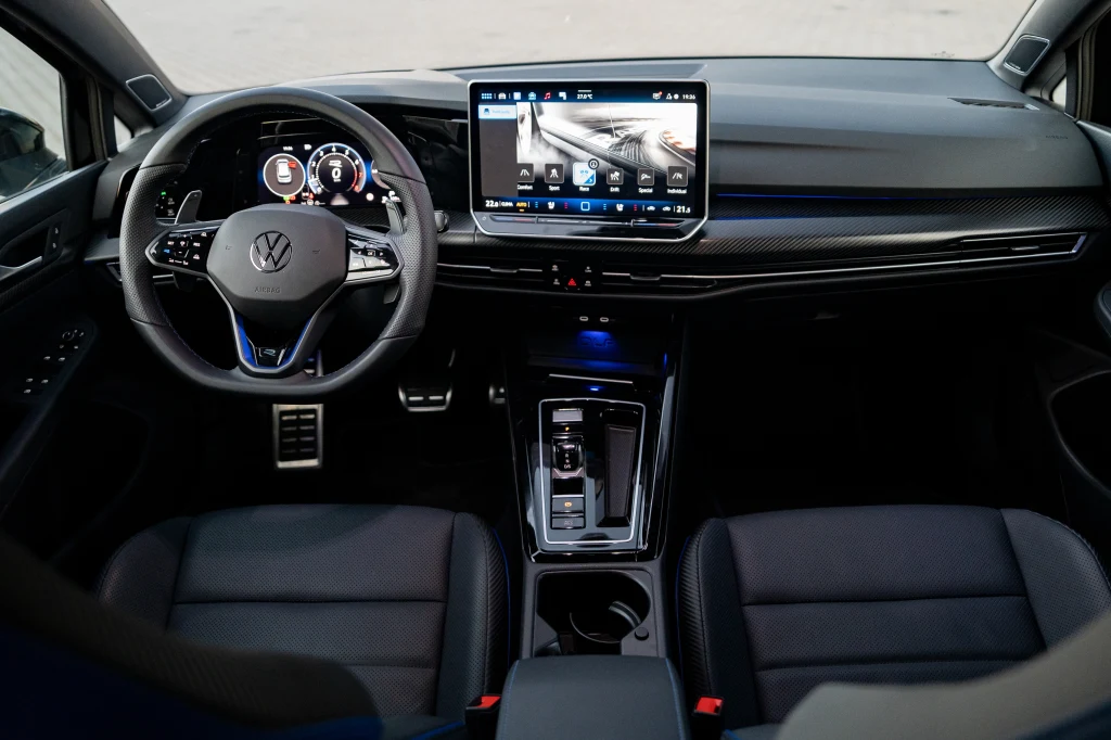 Wnętrze Golfa R jest jednolicie czarne, ale wysokiej jakości opcjonalna skóra nadaje mu nieco szlachetności Kokpit nowoczesnego samochodu marki Volkswagen z dużym ekranem dotykowym na konsoli centralnej, cyfrowymi zegarami i eleganckim wykończeniem wnętrza, fotele z czarnej skóry, widoczna dźwignia automatycznej skrzyni biegów.