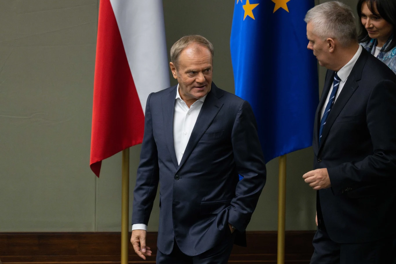 Premier Donald Tusk Mężczyzna w garniturze idzie obok innego mężczyzny, w tle widoczne są flagi Polski i Unii Europejskiej, atmosfera oficjalna, polityczna.
