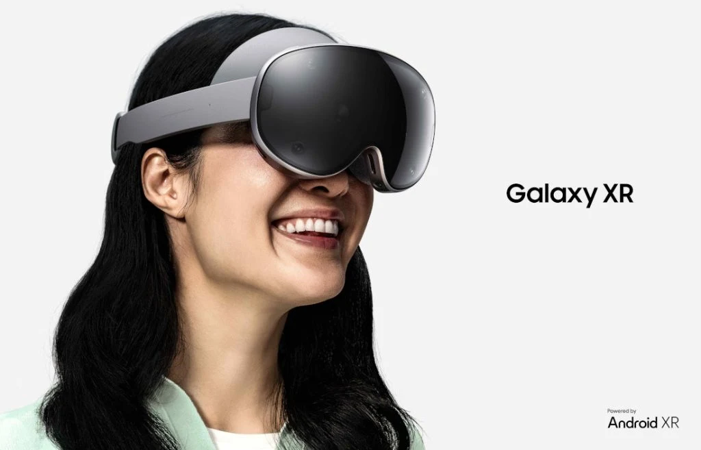 Kobieta o ciemnych włosach z uśmiechem na twarzy nosi na głowie nowoczesne gogle Galaxy XR, które służą do rozszerzonej rzeczywistości. Na białym tle widoczny jest napis Galaxy XR, a cała scena podkreśla nowoczesność i innowacyjność produktu.