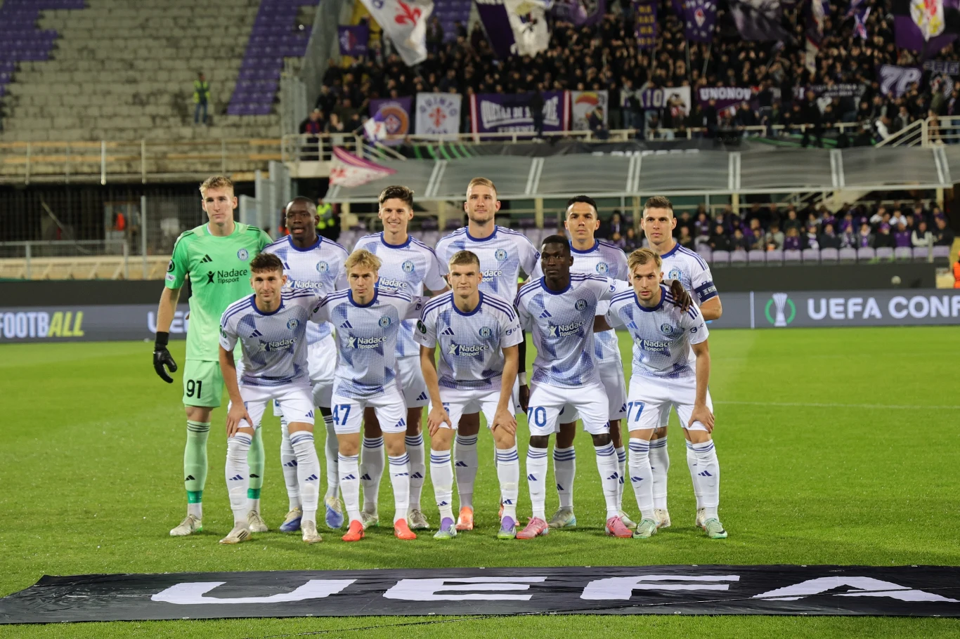 Grupa piłkarzy ustawionych w dwóch rzędach na murawie stadionu przed rozpoczęciem meczu, piłkarze ubrani w białe stroje z numerami, bramkarz w jasnozielonym uniformie, za nimi trybuny z kibicami i flagi klubowe.
