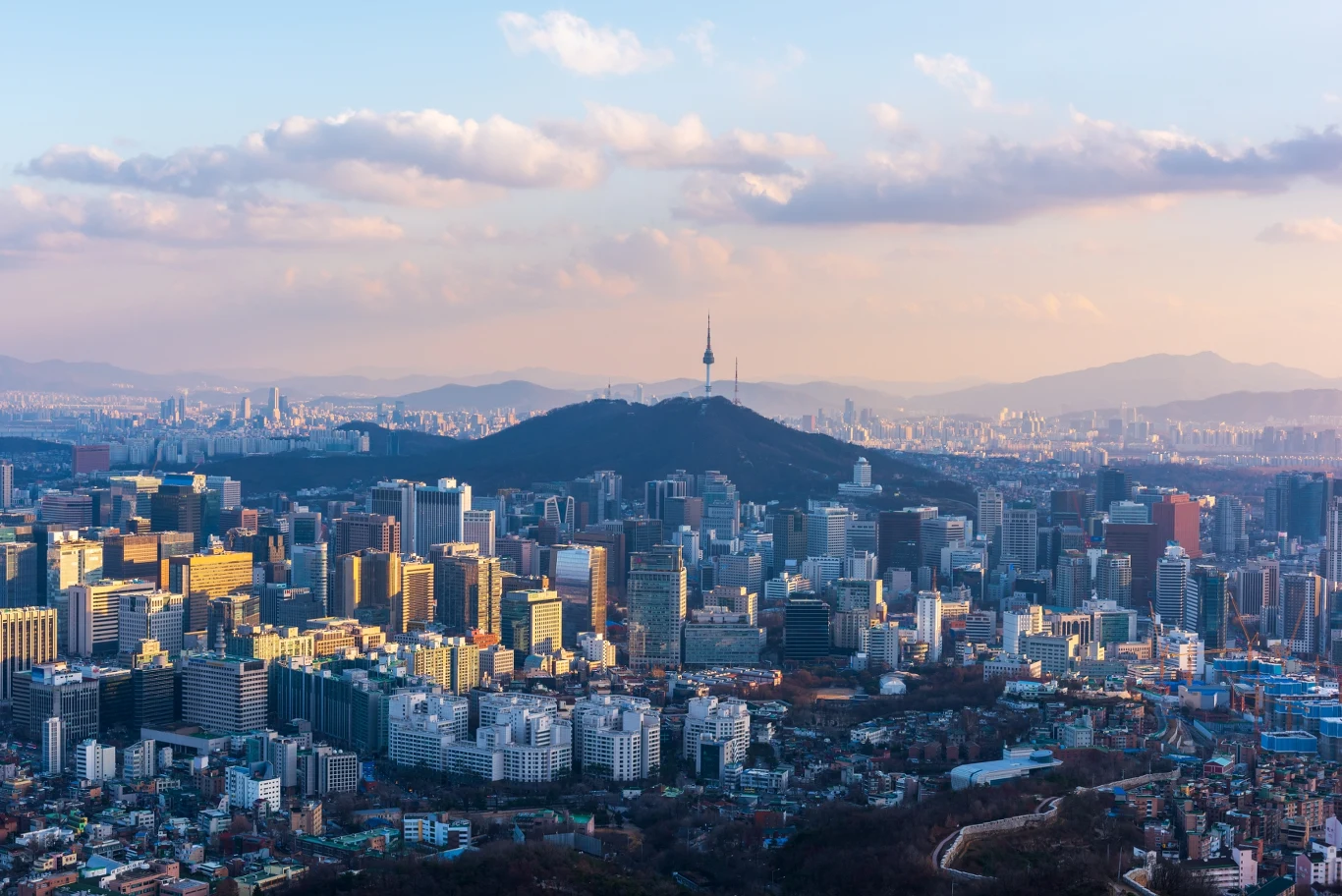 Loty do Korei Południowej znajdziemy już od 1800 zł w dwie strony Panorama nowoczesnych wieżowców miasta z charakterystycznym wzgórzem i wieżą telewizyjną Namsan na środkowym planie, w tle rozciągająca się dalsza część miasta i niebo z chmurami.