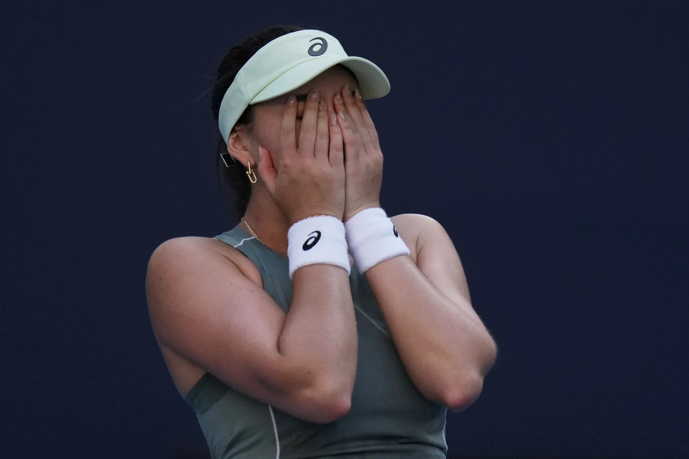 Niemiecka tenisistka Eva Lys Młoda kobieta ubrana w sportowy strój tenisowy zakrywa twarz obiema dłońmi, stojąc na ciemnym tle. Na rękach opaski sportowe, na głowie jasna czapka z daszkiem. Całość sylwetki wyraża silne emocje.