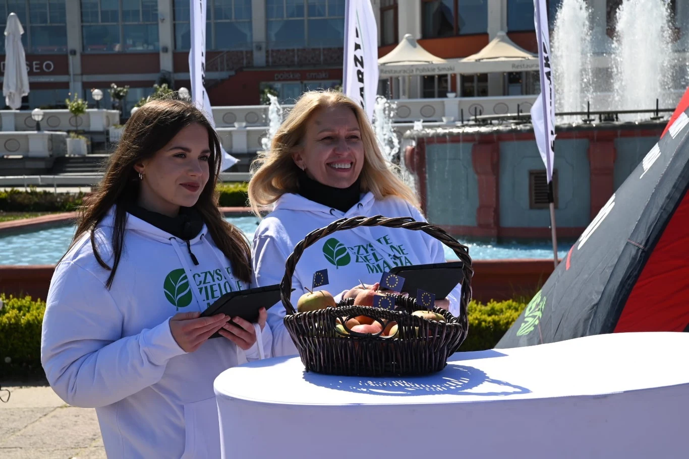 "Czyste, Zielone Miasta" w Sopocie Dwie kobiety w białych bluzach z nadrukiem promującym ekologię stoją przy stoliku na świeżym powietrzu, przed nimi wiklinowy kosz z owocami, w tle fontanna i namiot eventowy.