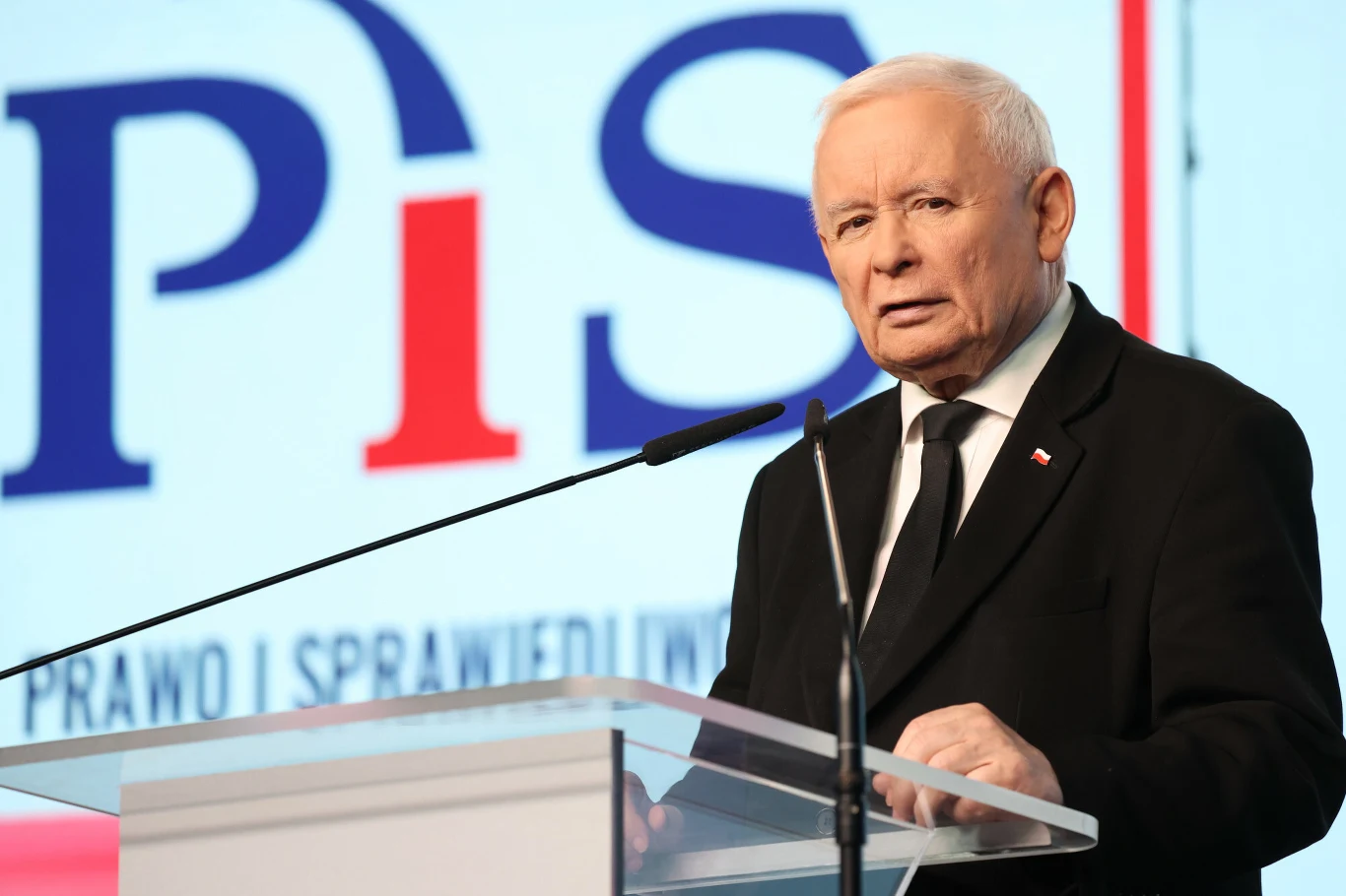 Jarosław Kaczyński, 15.10.2025 r. Starszy mężczyzna w ciemnym garniturze i białej koszuli stoi za przezroczystą mównicą, mówi do mikrofonu na tle logo i napisu partii politycznej Prawo i Sprawiedliwość.