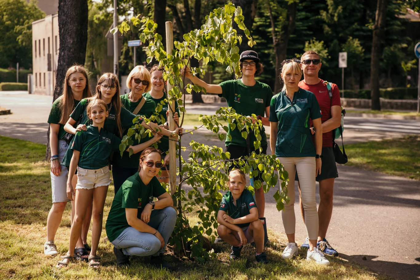 "Czyste, Zielone Miasta" w Lesznie - sadzenie lip Grupa ludzi w zielonych koszulkach pozuje wokół świeżo posadzonego drzewa w parku miejskim, kilka osób klęczy przy drzewku, pozostali stoją, wszyscy uśmiechnięci, za nimi rozciąga się aleja drzew i budynki.