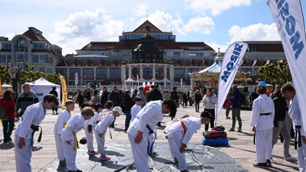 Grupa dzieci i młodzieży w strojach do karate wykonuje ukłon na macie ustawionej na otwartym placu miejskim, w tle liczne osoby spacerujące oraz charakterystyczny budynek uzdrowiskowy.