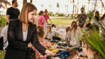 Dziennikarka przeprowadzająca wywiad z kobietą podczas wydarzenia plenerowego dla dzieci, w tle grupa dzieci i dorosłych biorących udział w warsztatach i zabawach przy stołach.