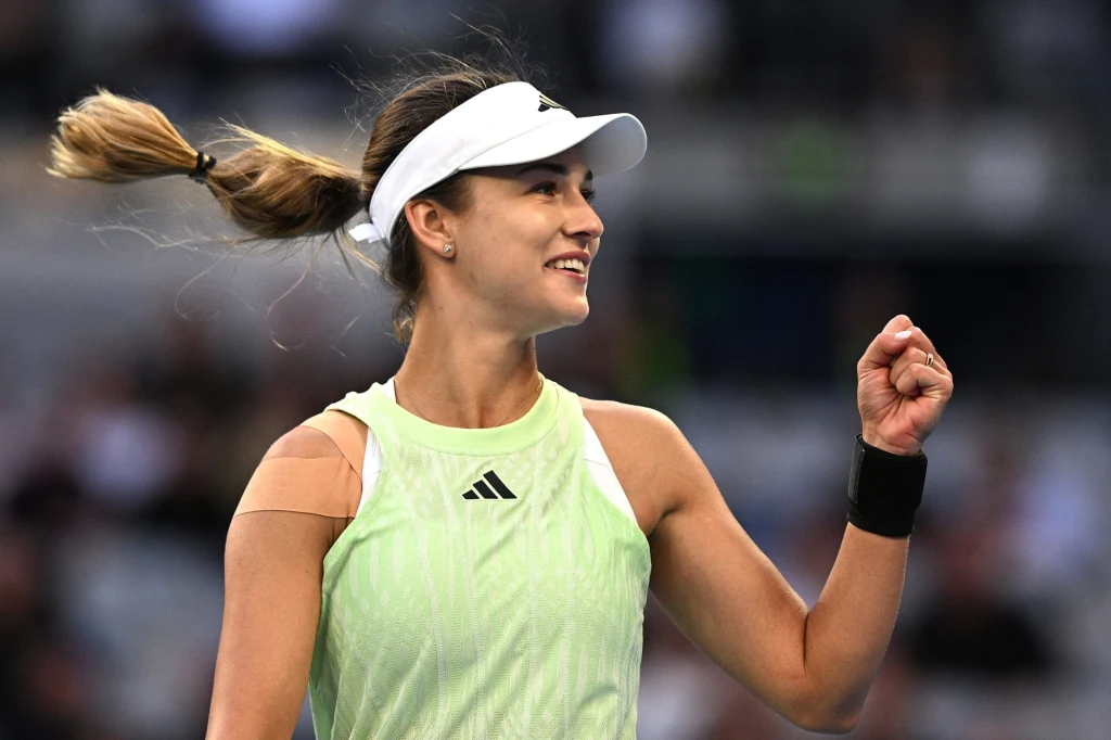 Tenisistka w jasnym stroju sportowym i białym daszku cieszy się po wygranym punkcie na korcie, unosząc zaciśniętą pięść w geście triumfu, w tle rozmyta publiczność.