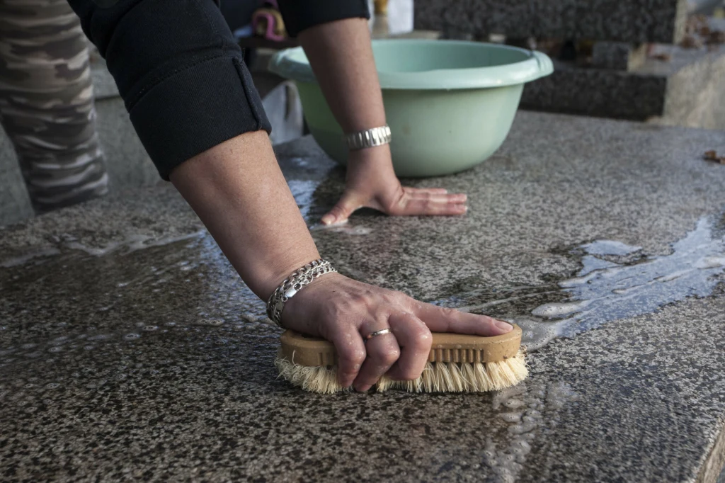 Ręka osoby w bransoletce czyszcząca szczotką marmurową powierzchnię, w tle plastikowa miska z wodą i fragment drugiej ręki opierającej się o blat.
