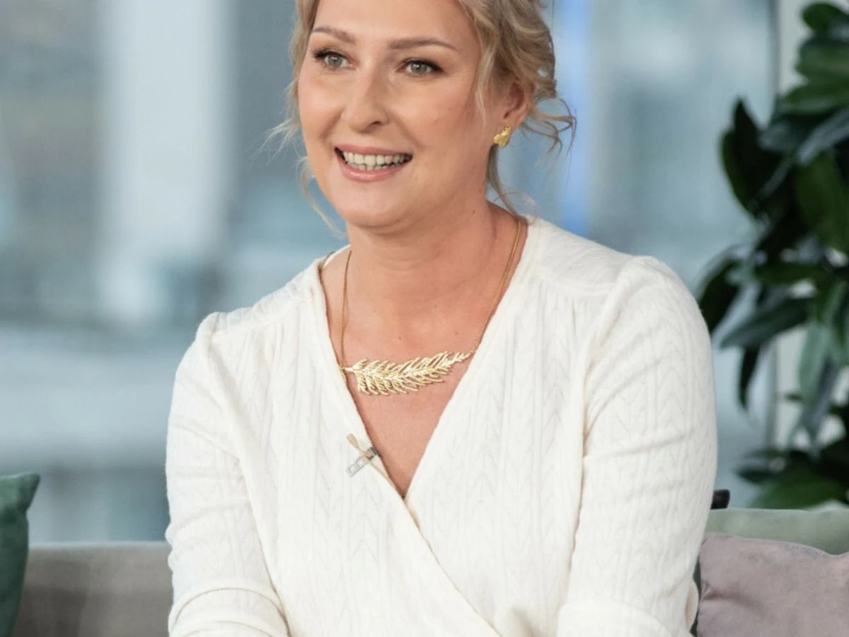 Anita Lipnicka Uśmiechnięta blondynka w białej bluzce i złotej biżuterii siedzi na sofie, w tle są rozmyte zielone rośliny i jasne wnętrze.