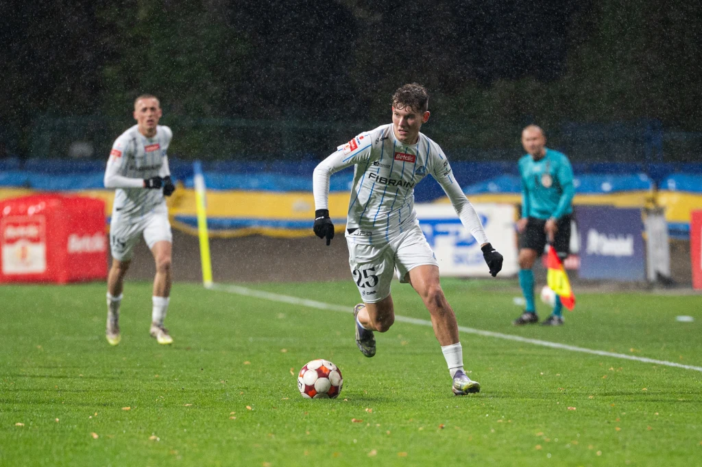 Piłkarze Stali Rzeszów w sezonie 2025/2026 I ligi Piłkarz w białym stroju prowadzi piłkę po mokrym boisku podczas meczu nocą, za nim biegnie inny zawodnik tej samej drużyny, a w tle widoczny jest sędzia oraz fragment trybun.