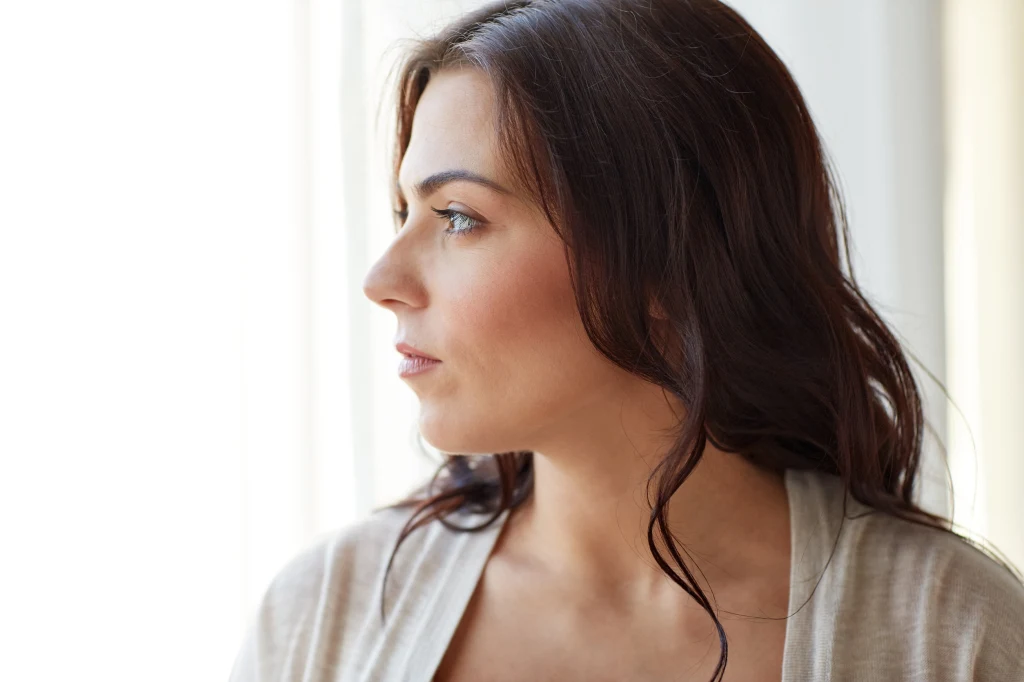 Panna odczuje planetarne napięcie Młoda kobieta o ciemnych włosach patrząca z zamyśleniem przez okno, światło słoneczne delikatnie oświetla jej twarz, widoczne są naturalne rysy twarzy oraz spokojny wyraz.