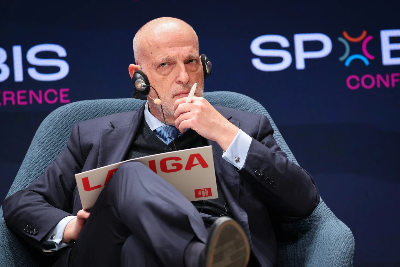 Łysy, starszy mężczyzna w garniturze siedzi w fotelu, trzyma w dłoni dokument z napisem 'LA LIGA', na głowie ma słuchawki z mikrofonem, skupiony wyraz twarzy wskazuje na rozważania lub udział w dyskusji podczas konferencji.