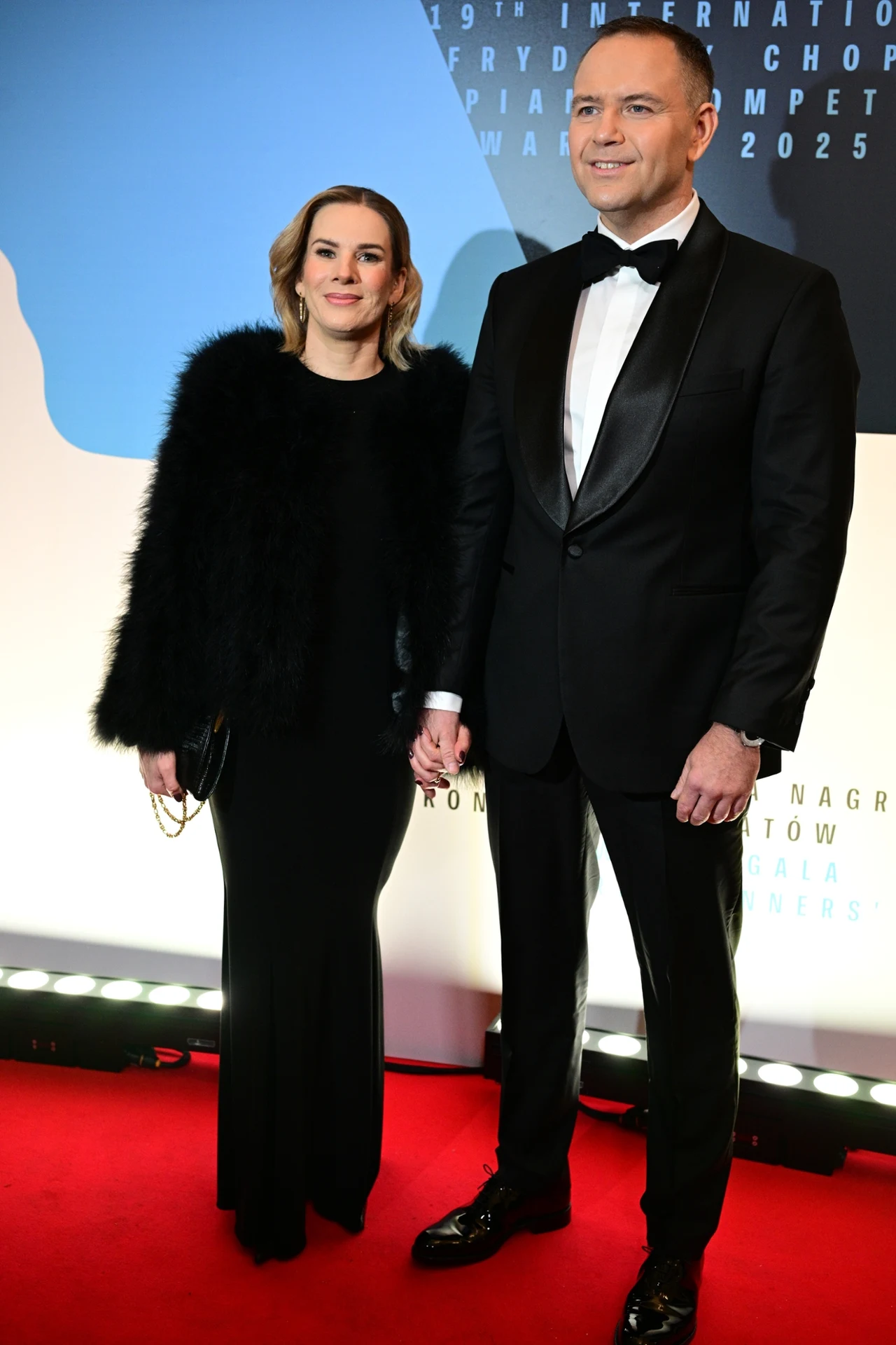 Marta i Karol Nawroccy Elegancka para trzymająca się za ręce pozuje na czerwonym dywanie; kobieta ubrana w czarną suknię i futerko, mężczyzna ma na sobie klasyczny smoking z muchą, w tle oficjalna ścianka wydarzenia.