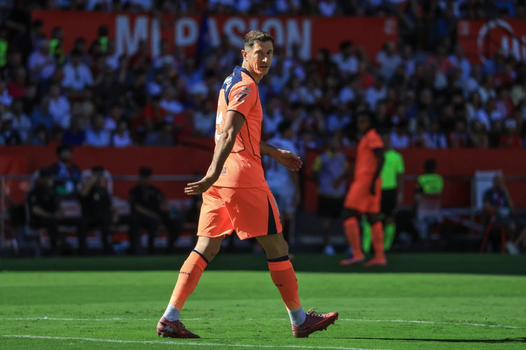 Piłkarz w pomarańczowym stroju sportowym idzie po murawie stadionu podczas meczu, w tle widać kibiców na trybunach oraz innych zawodników.