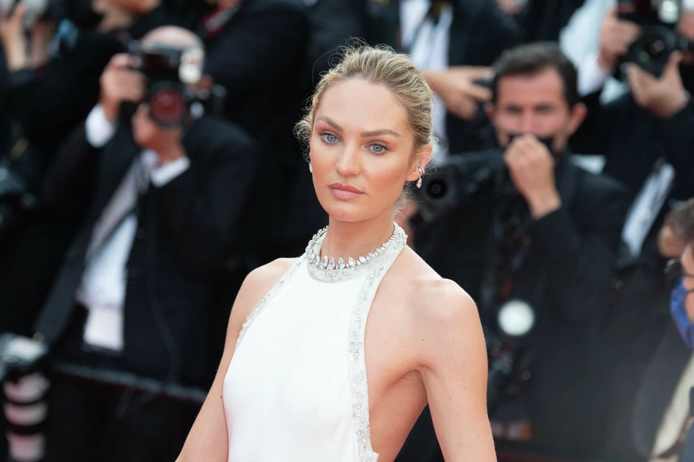 Candice Swanepoel skończyła 37 lat Elegancko ubrana kobieta z blond włosami założonymi gładko do tyłu, w białej, wyciętej sukni z ozdobnym naszyjnikiem, stoi na tle tłumu fotografów i reporterów na czerwonym dywanie.