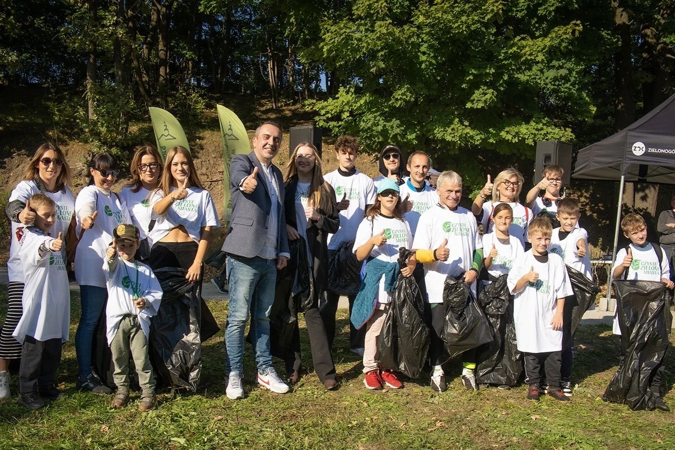 "Czyste, Zielone Miasta" w ZIelonej Górze Grupa osób w różnym wieku ubrana w białe koszulki bierze udział w akcji sprzątania terenów zielonych, trzymając worki na śmieci i pokazując gesty aprobaty na tle drzew.