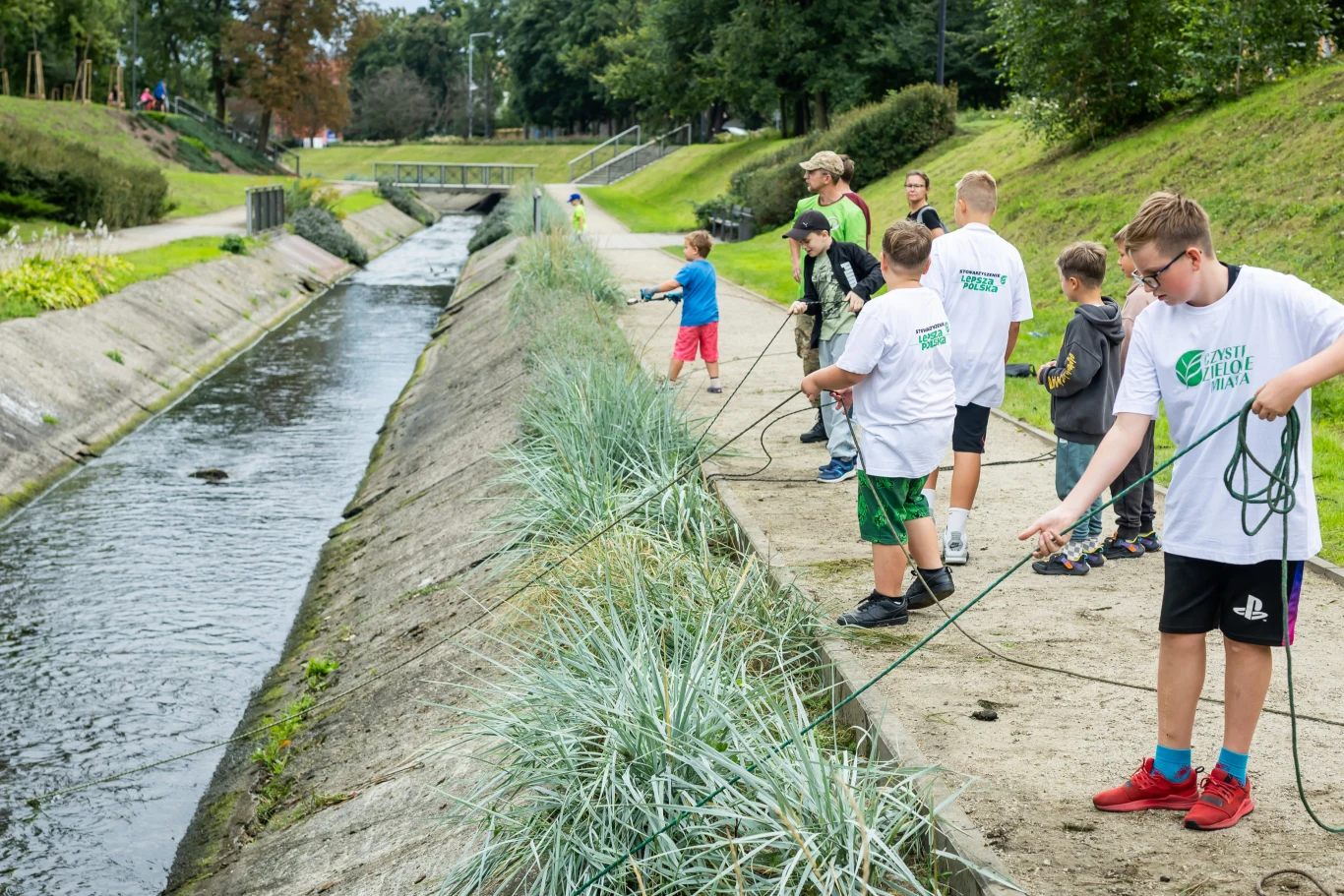 "Czyste, Zielone Miasta" w Bydgoszczy - magnesowanie odpadów Grupa dzieci i dorosłych stojących przy betonowym kanale rzeki, trzymających liny, zaangażowanych w wspólne zajęcia na świeżym powietrzu, otoczonych zielenią, w spokojnej okolicy miejskiej.