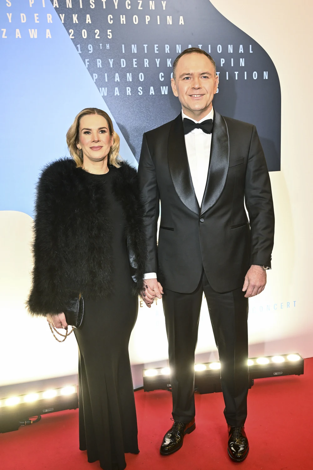 Podczas gali wręczenia nagród Marta Nawrocka zachwyciła wytworną suknią  Elegancko ubrana para stoi na czerwonym dywanie, kobieta w długiej czarnej sukni i futrzanej narzutce, mężczyzna w smokingu z muchą, oboje uśmiechnięci, w tle ścianka promująca Konkurs Chopinowski.
